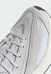Zapato deportivo gris claro que combina materiales de ante y malla. Diseño de olas distintivo, con cordones blancos y acentos texturizados.