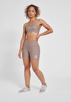 Jonge vrouw staat met haar hand op haar heup, draagt een taupekleurige sportbeha, bijpassende shorts met hoge taille en grijze sneakers tegen een witte achtergrond.