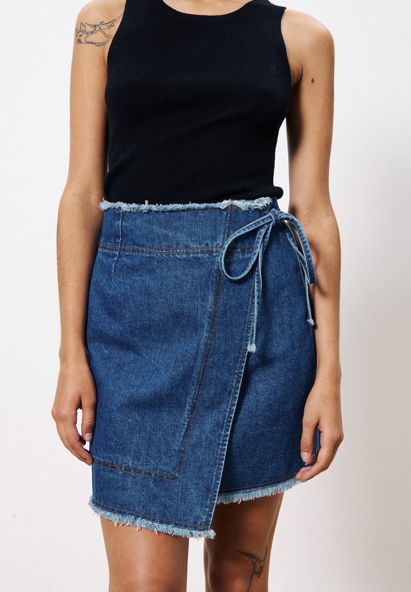 Jupe en denim avec un ourlet effiloché, couleur bleue, ceinture réglable à la taille et détails de couture visibles. Associée à un débardeur noir.