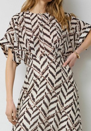 Žena u krem haljini kratkih rukava s crnim cik-cak uzorcima, prednjim patentnim zatvaračem i zlatnom narukvicom na desnom zglobu.