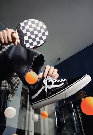 Una persona hace malabares con pelotas de ping-pong naranjas y blancas usando una pala a cuadros mientras se ata una zapatilla negra.