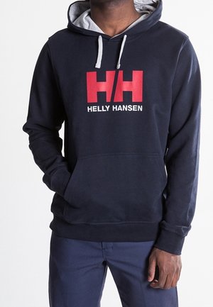 Personne portant un sweat à capuche Helly Hansen bleu marine avec des cordons blancs et un logo HH rouge, associé à un pantalon bleu foncé.