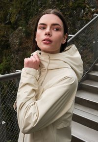 Woman wearing a beige hooded jacket, holding the collar, standing next to metal stairs and a safety railing outdoors near rocky terrain.