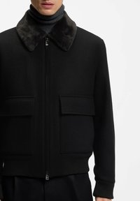 Mann trägt eine schwarze Reißverschlussjacke mit großen Klappentaschen und einem dunklen Kunstpelzkragen über einem schwarzen Rollkragenpullover.