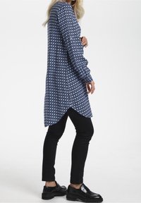 Tunika z długim rękawem w kolorze granatowym z geometrycznym wzorem, z zaokrąglonym dołem i detalem guzika na rękawie, w zestawie z czarnymi dopasowanymi spodniami.