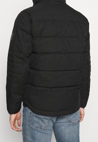 Mann in schwarzer gepolsterter Jacke und blauen Jeans, von hinten gegen einen schlichten hellen Hintergrund gezeigt.