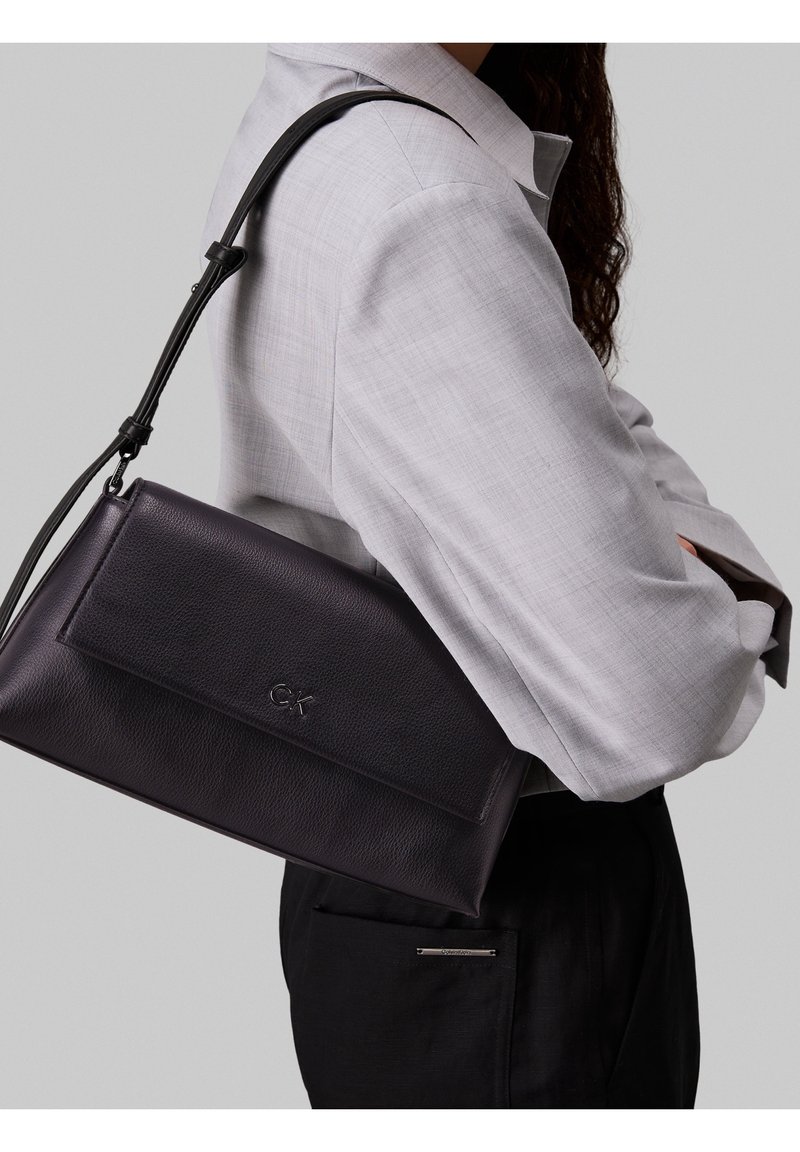 Femme portant un blazer gris clair et un pantalon noir, portant un sac à épaule en cuir noir Calvin Klein sur son côté droit.
