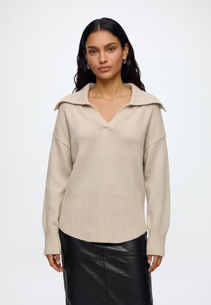 Kvinde med langt mørkt hår iført beige striktrøje med bred krave og sort lædernederdel, stående mod en ensfarvet lys baggrund.