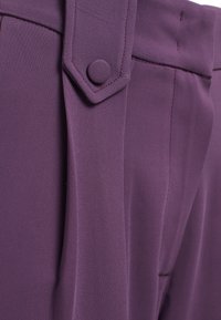 Pantalons en tissu texturé violet avec un pli marqué, des plis latéraux et une taille sécurisée par un bouton. Les détails incluent des coutures ajustées.
