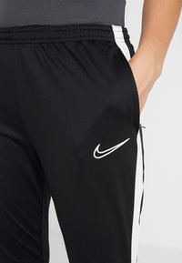 Nahaufnahme einer Hand, die in der Tasche einer schwarzen Sporthose mit weißen Seitenstreifen und dem weißen Nike-Logo auf dem Oberschenkel steckt.