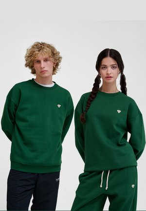 Green cotton sweatshirts with relaxed fit and ribbed cuffs, featuring a small embroidered heart logo on the chest. Styled with matching bottoms.