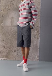 Graues Langarm-Polo mit roten Streifen, kombiniert mit dunkelgrauen Shorts. Weiße Sneakers und leuchtend rote Socken vervollständigen das Outfit.