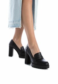 Zapatos de tacón alto negros con una plataforma gruesa y punta redonda, que cuentan con un detalle de correa y suela texturizada, combinados con una falda de mezclilla clara.