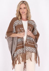 Brauner und grauer Poncho mit geometrischem Muster, mit Fransen und weicher Textur. Getragen über einem weißen Oberteil.