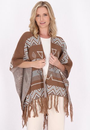 Ruskea ja harmaa kuviollinen poncho, jossa on geometrinen malli, reunassa fringejä ja pehmeä tekstuuri. Pukeutuneena valkoisen paidan päälle.