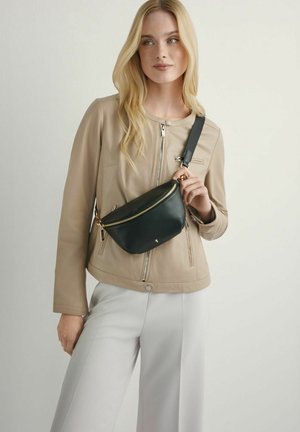 Kobieta ubrana w beżową kurtkę i białe spodnie, trzymająca czarną torebkę na pasku z złotym zamkiem błyskawicznym na jednolitym tle.