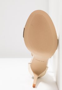 Chaussure à talon haut beige inclinée vers le haut avec semelle texturée visible et nom de la marque "ALDO" sur la voûte plantaire sur fond blanc.