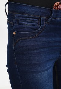 Los jeans de mezclilla azul oscuro presentan un corte ajustado, un bolsillo frontal con detalles de costura y un botón de cobre como acento cerca de la cintura.