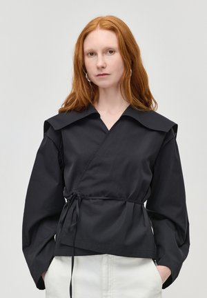 Žena s červenými vlasmi, oblečená v čiernej viazanej blúzke so širokým golierom a bielych nohaviciach, stojaca s rukami v vreckách na jednoduchom pozadí.