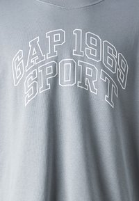 Šviesiai pilkas medvilninis džemperis su baltais blokiniais užrašais "GAP 1969 SPORT" krūtinės srityje, turintis klasikinį apvalų kaklą.
