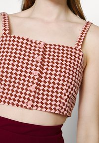 Femme portant un crop top à motifs rose et marron avec de fines bretelles et cinq boutons à l'avant, associé à un pantalon taille haute de couleur rouge foncé.