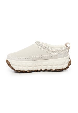 Zapatos tipo slip-on en beige claro con un upper texturizado, suela acolchada y una suela de goma marrón para tracción. Costuras decorativas alrededor del cuello.