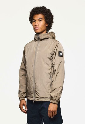 Mann mit lockigem Haar, der eine hellbraune Kapuzenwindjacke und dunkle Hosen trägt, im Stehen vor einem schlichten hellen Hintergrund.