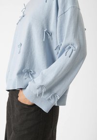Light blue sweater with decorative bows on the sleeves, paired with dark pants, shown from the side with a hand in the pocket.