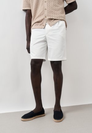 Férfi, aki bézsszínű rövid ujjú kötött inget, fehér térdig érő rövidnadrágot és fekete bebújós cipőt visel, fehér háttér előtt áll.