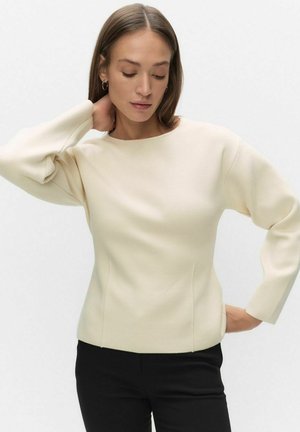 Vrouw met lang bruin haar, gekleed in een crèmekleurige top met lange mouwen en zwarte broek, poseert met een hand achter haar nek en haar ogen gesloten.