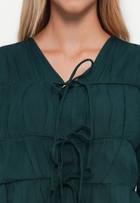 Kvinne med mørkegrønn bluse med V-hals og tre knyttede sløyfer nedover fronten, som viser øvre bryst og blond hår.