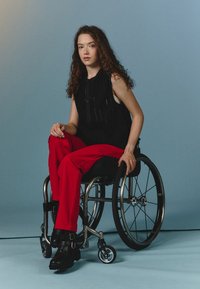 A person in a black sleeveless top and red pants sits in a wheelchair. The wheelchair features metallic accents and sleek design.