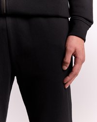 Pravá ruka spočívajúca na boku čiernych nohavíc, viditeľná časť rukáva čiernej mikiny so zipsom, svetlé pozadie.