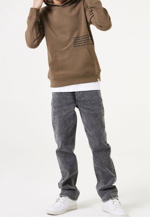 Sweat à capuche marron avec une poche frontale et un détail texte sur le côté, associé à un jean gris clair et des baskets blanches. Texture de tissu lisse.