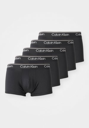 Pięć czarnych bokserów Calvin Klein ułożonych w stos po przekątnej z białą gumką z logo na jednolitym białym tle.