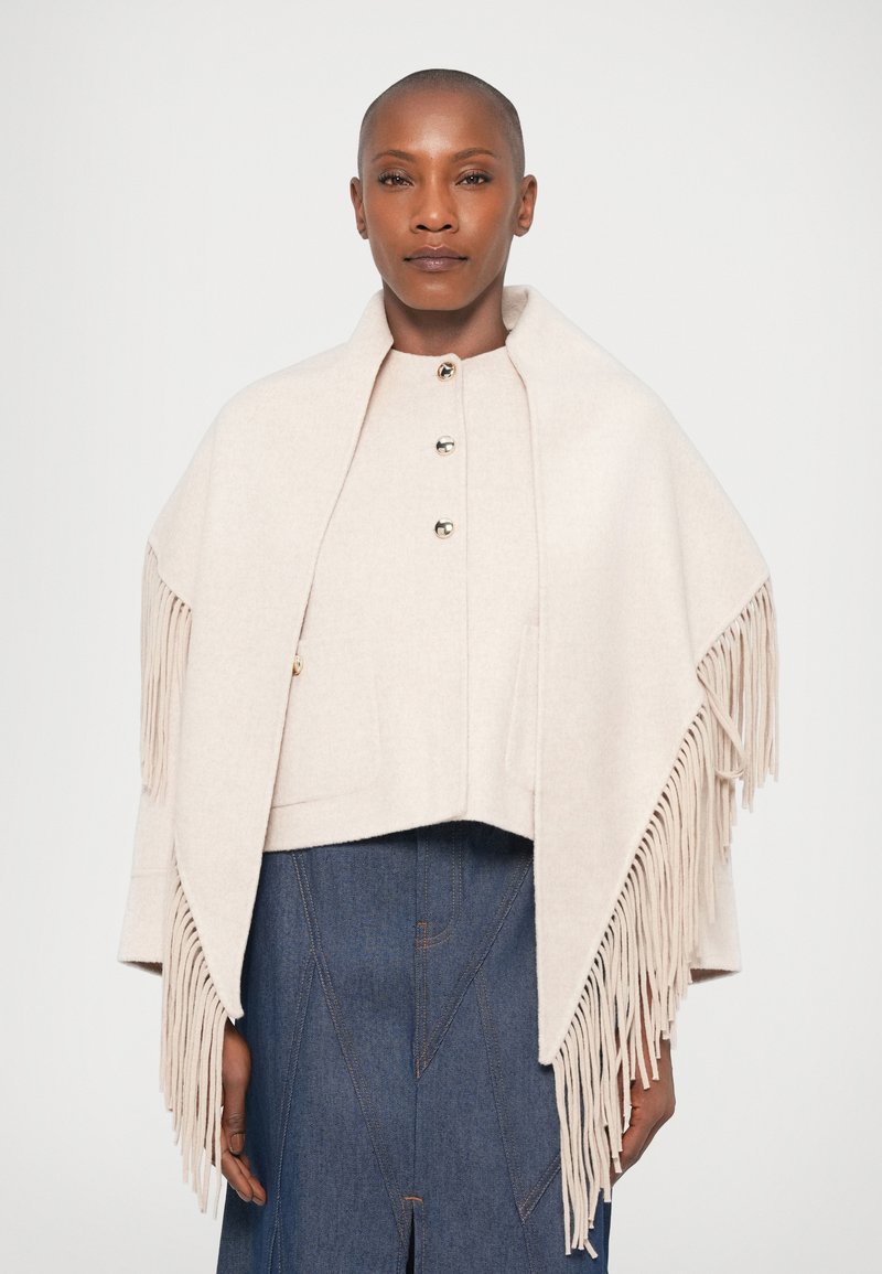 Ljusbeige fransad poncho i mjukt tyg, med rund hals och knappstängning, bärs över en blå denimkjol.