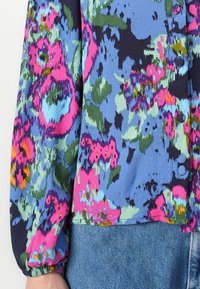 Blouse florale avec un fond bleu vif et des motifs floraux roses, verts et marine, des manches longues bouffantes et des fermetures à boutons.