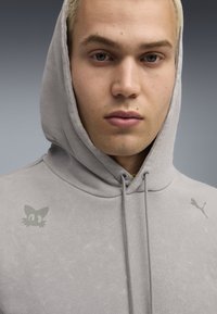 Grå hættetrøje med en frontlomme, justerbar snøre, med et lille kattegrafik og et Puma-logo på skulderen. Blød tekstur.