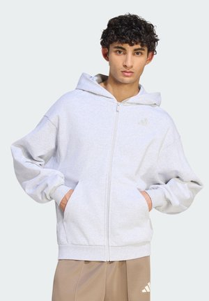 Ung mand med krøllet hår iført en lysegrå lynlåshoodie og beige bukser, hænderne i lommerne, mod en ensfarvet baggrund.