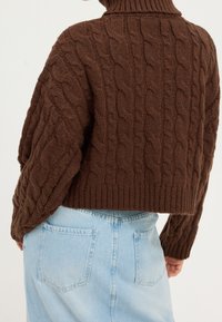 Hnědý svetr s pleteným vzorem a vysokým límcem, s dlouhými rukávy a zkrácenou délkou. Nosený přes světle modrou džínsovou sukni s kapsami na zadní straně.