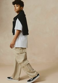 T-shirt blanc léger, pantalon cargo beige avec de multiples poches, pull noir noué autour des épaules et baskets noires.