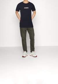 Marineblaue T-Shirt mit kurzen Ärmeln und "GUESS"-Logo, olivgrüne Hose und weiße Sneakers mit roten Akzenten. Einheitlicher Hintergrund.