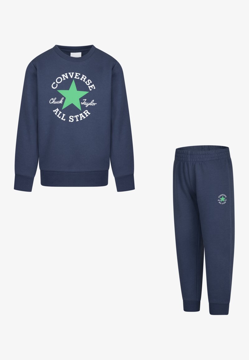 Tumši zils džemperis ar baltu un zaļu "Converse All Star" grafiku; kombinēts ar atbilstošām biksēm, kuru sānos ir mazs logo.