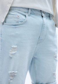 Ljust blå slitna jeans med flera repas, fem fickor, silverfärgade detaljer och en något urblekt textur.