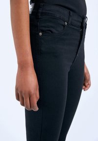 Black jeans with a slim fit, featuring a zip fly, front pockets, and a smooth texture. Metal hardware accents enhance the design.