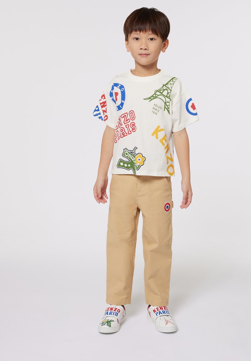 T-shirt graphique blanc avec des imprimés colorés, pantalon beige et des chaussures slip-on à motifs arborant des logos. Enfant se tient de manière neutre sur un fond blanc.
