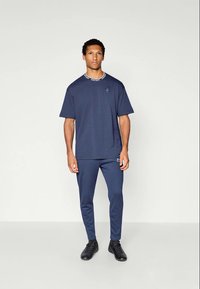 Mand står mod en ensfarvet baggrund iført en marineblå t-shirt, matchende slim-fit bukser og sorte sneakers, vendt fremad med en neutral ansigtsudtryk.