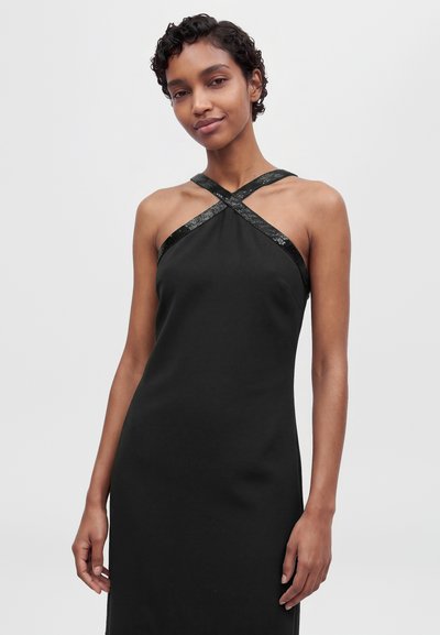 Modèle aux cheveux courts et bouclés, portant une robe noire sans manches à bretelles croisées, devant un fond uni clair.