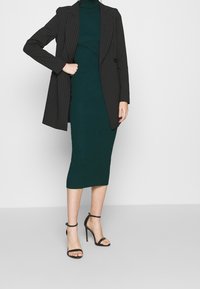 Blazer noir à rayures fines porté sur une robe midi côtelée vert foncé. Mannequin portant des sandales à talons hauts noires à lanières. Textures lisses et silhouette ajustée.