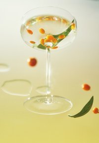 Glass coupe fylt med klar væske, oransje sfærer og grønt blad. Reflekterende overflate nedenfor, myk gul bakgrunn.